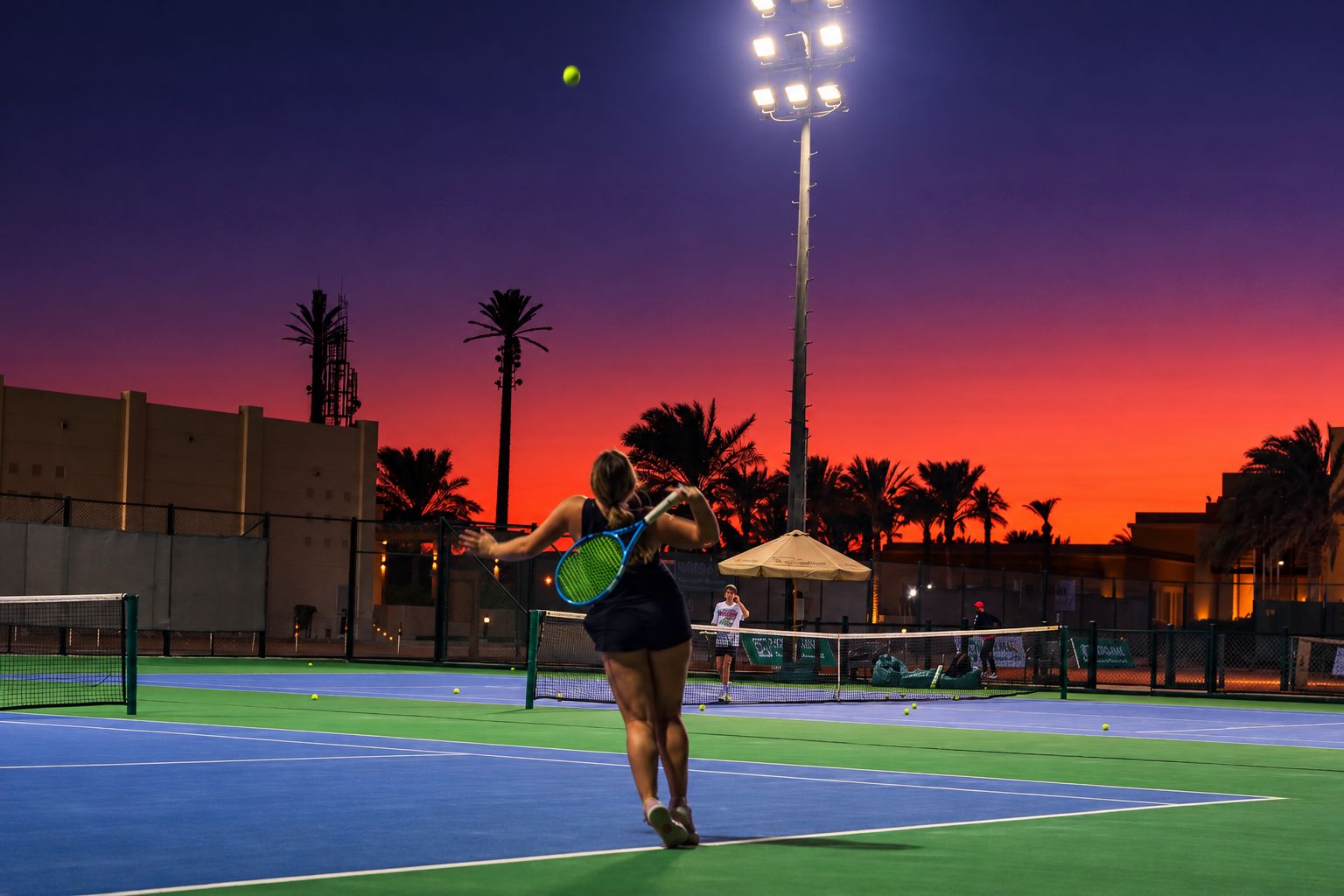 A player serving under floodlights at sunset at El Gouna Tennis Club, palms behind the courts